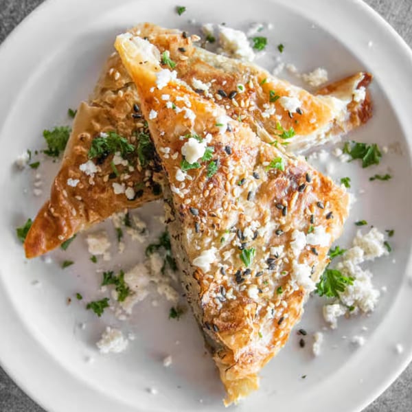 Two golden-brown Greek spanakopita pastries, topped with feta, sesame seeds, and parsley, on a white plate.